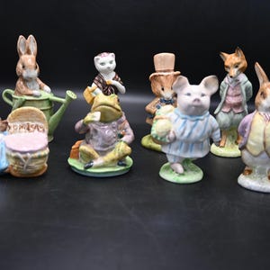 Może przedstawiać: Kolekcja ceramicznych figurek przedstawiających antropomorficzne zwierzęta. Figurki obejmują królika, kota, żabę, świnię i lisa, każdy ubrany w szczegółowe ubrania. Figurki są pomalowane w różnych kolorach i stoją na małych, zielonych podstawach.