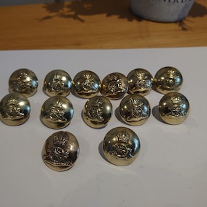 Może przedstawiać: Kolekcja dwunastu złotych metalowych guzików. Każdy guzik ma szczegółowy wytłoczony wzór, w tym koronę i armatę. Guziki są okrągłe i mają błyszczące wykończenie, co sugeruje, że są zabytkowe lub antyczne.