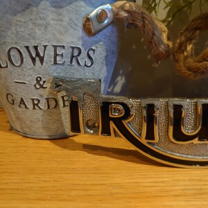 Puede incluir: Un cubo de metal galvanizado con las palabras "FLOWERS & GARDEN" en relieve en el lateral. Un letrero decorativo de metal con la palabra "TRIUM" est&aacute; delante del cubo. Un asa de cuerda est&aacute; sujeta al cubo.