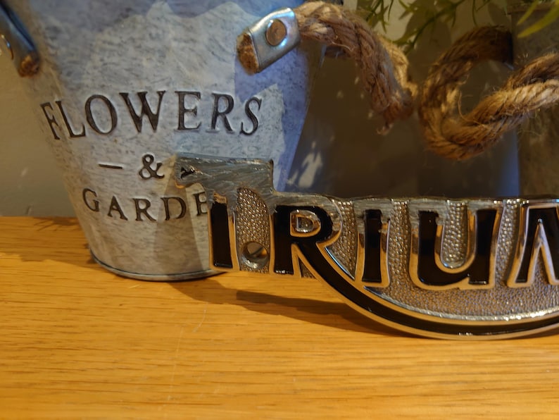 Puede incluir: Un cubo de metal galvanizado con las palabras "FLOWERS & GARDEN" en relieve en el lateral. Un letrero decorativo de metal con la palabra "TRIUM" est&aacute; delante del cubo. Un asa de cuerda est&aacute; sujeta al cubo.