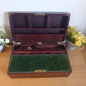 Op de afbeelding: Een open, antieke houten schrijfkist met een donkere afwerking, met compartimenten, een groen vilt schrijfoppervlak en messing hardware. De kist staat op een houten tafel, met kleine bloemen in potten aan weerszijden.