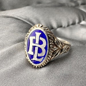 Puede incluir: Un anillo de plata con un óvalo de esmalte azul que presenta las iniciales "LB" en blanco. El anillo tiene un diseño floral detallado en la banda.