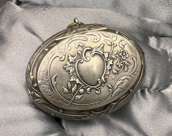 May include: A silver locket with an ornate floral design and a heart-shaped center. The locket has a hinged lid and a loop for attaching a chain.