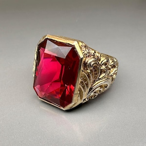 Peut inclure: Une bague en or avec un motif orné et une grande pierre précieuse rectangulaire rouge.