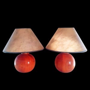 May include: Two matching table lamps. Each lamp has a round, red base and a beige lampshade with a dark gray trim. The lampshades are conical in shape. The background is black.