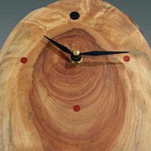 May include: A wooden clock with a natural wood grain finish. The clock face has black hands and red and black markers. The clock is mounted on a round piece of wood.