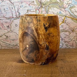 Puede incluir: Un vaso de madera hecho a mano con una estética natural y orgánica. El vaso tiene una forma cilíndrica con una abertura ancha y un acabado liso y pulido. Los patrones de la veta de la madera crean un aspecto único y rústico. El vaso se exhibe sobre una superficie de madera.