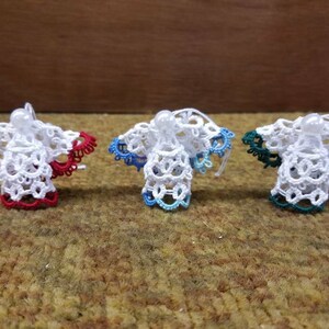May include: Three white crocheted angel ornaments with red, blue, and green accents. The ornaments are hanging from a string.