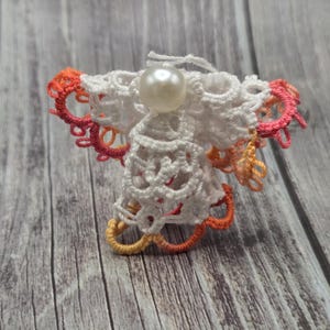 May include: A white crocheted angel ornament with a pearl bead in the center. The angel has pink and orange accents around the edges.