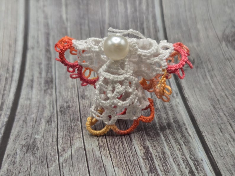 May include: A white crocheted angel ornament with a pearl bead in the center. The angel has pink and orange accents around the edges.