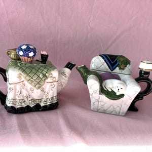 May include: Two ceramic teapots shaped like miniature furniture. The left teapot resembles a table with a green checkered cloth, a blue basket, and a black handle. The right teapot is shaped like a white armchair with a sleeping cat and a lamp.