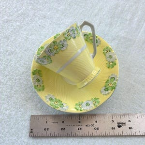 May include: A yellow teacup and saucer set featuring a raised white and green floral pattern and a ribbed texture. The saucer measures approximately 5.5 inches in diameter, and the set is positioned next to a metal ruler for scale.
