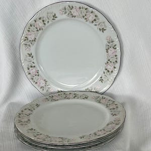 May include: A set of white porcelain plates with a floral rose design. The plates have a scalloped edge and a black trim. The roses are pink and white with green leaves. The set includes four plates.