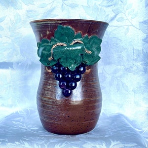 May include: A brown ceramic vase with a grapevine design. The vase has a textured surface and a wide mouth. The grapevine design is three-dimensional and features green leaves and purple grapes.