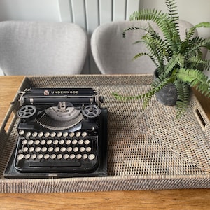 May include: A black Underwood typewriter with white keys is on a woven tray, alongside a potted fern. The rectangular tray has handles and is on a wooden table. Two grey chairs are in the background.