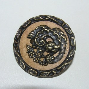 May include: A round, wooden button with a black metal rim and a detailed, embossed design of a mythical creature with wings and a floral motif.