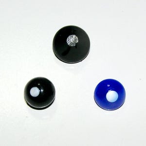 May include: Three circular cabochons: one large black piece with a textured silver center, one small black piece with a white dot, and one small blue piece with a white dot. These items are smooth, rounded, and feature distinct color patterns.