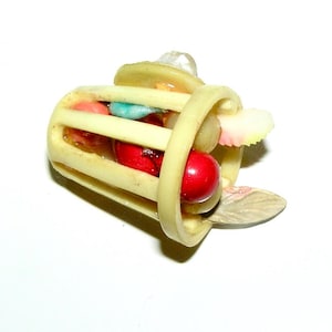 May include: A small, cream-colored, cylindrical cage-like object with vertical slats. Inside the structure are several small, colorful, spherical and leaf-shaped decorative elements in shades of red, pink, blue, and light green.
