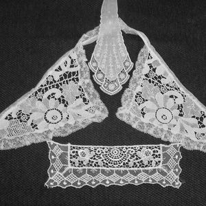 May include: White lace collar and cuffs set. The set includes a triangular collar, two matching cuffs, and a rectangular piece. The lace features floral and geometric patterns, with scalloped edges. The pieces are arranged on a dark background.