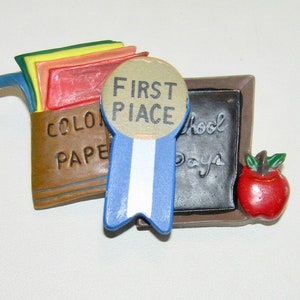 Puede incluir: Una caja marrón con las palabras "Color Paper" en ella, una cinta azul con un círculo dorado que dice "First Place", una pequeña pizarra con las palabras "School Days" en ella, y una manzana roja.