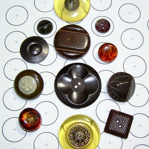 May include: A selection of antique buttons in diverse designs and hues. The assortment features a yellow button with a coiled centre, a dark brown, flower-shaped button, and a square button with diagonal markings. The buttons are displayed on a white backdrop with circular patterns.