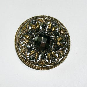 May include: A circular brooch featuring an antique gold-toned metal frame with an intricate openwork leaf pattern. The center is set with a large, faceted dark gray stone, surrounded by smaller, round, faceted dark gray beads throughout.