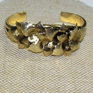 May include: A gold-toned bracelet with a design of leaves. The bracelet is open and has a simple, elegant design.