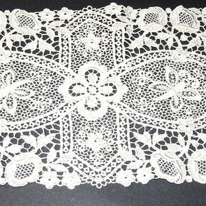 May include: An off-white, rectangular lace doily with an intricate floral and geometric design. The lace features oval and square patterns, with detailed flower and leaf motifs throughout. The edges are scalloped, adding a delicate finish to this decorative textile.