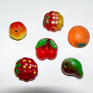 Op de afbeelding: Een verzameling van zes fruitvormige kralen, waaronder een gele en rode perzik, een rode en gele framboos, een textuurrijke sinaasappel, een paar rode kersen met groene blaadjes, een rode bes met gele stippen en een groen-bruine peervormige kraal.