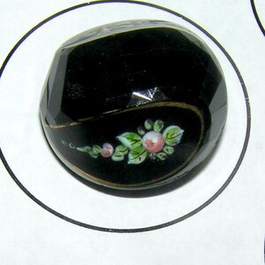 May include: A faceted, black button with a floral design. The button has a gold trim and a painted design of pink roses and green leaves. The button is round and has a glossy finish.