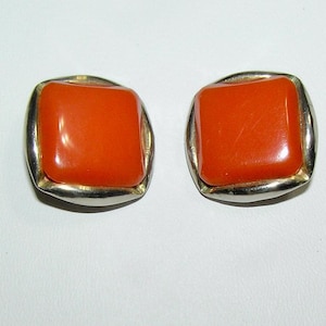 May include: A pair of square-shaped stud earrings. Each earring features a vibrant orange, glossy, square stone set within a silver-toned metal frame. The earrings are displayed against a white background, showcasing their simple yet elegant design.