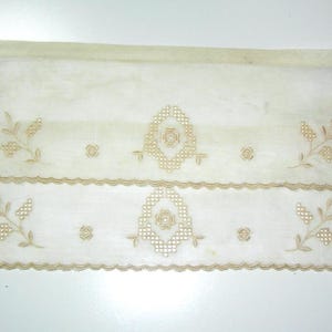 May include: Cream-colored, sheer fabric with embroidered floral and geometric designs. The fabric has a scalloped edge and is likely a decorative element for a window or other home decor item.