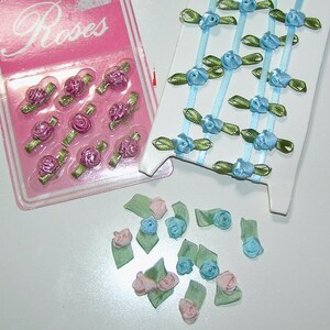 May include: A collection of decorative floral embellishments. The image shows pink and blue ribbon roses, some packaged on a pink card with the word "Roses" and others loose. The roses have green leaves and are suitable for crafts.