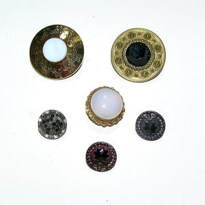 May include: A collection of six circular buttons featuring varied designs. Two large buttons have metallic bases with white and black centers. A central button has a white dome. Three smaller buttons feature dark, textured, and star-shaped patterns.