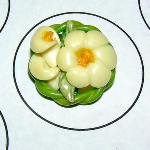 May include: A floral-themed brooch featuring a cream-colored flower with an orange center and two mushroom-shaped elements. The flower and mushrooms are set within a green, woven-like border. The brooch is on a white background with a black circle.