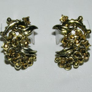 Peut inclure: Une paire de boucles d'oreilles à clip dorées vintage. Chaque boucle d'oreille présente un motif floral avec des strass couleur ambre et des accents de feuilles. Les boucles sont montées sur du carton blanc.
