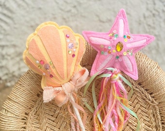 Puede incluir: Dos varitas de fieltro con una concha de color melocotón y una estrella de mar rosa. La concha está adornada con lentejuelas y una cinta color melocotón. La estrella de mar rosa presenta una gema central color ámbar, lentejuelas y un mango decorado con hilos y cintas de colores.