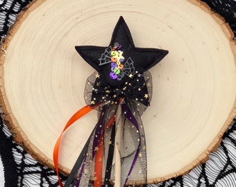 May include: A black felt star wand featuring a central cluster of colorful sequins and spiderweb accents. The wand includes a black tulle bow with gold star patterns and a collection of hanging ribbons in orange, purple, black, and gold glitter.