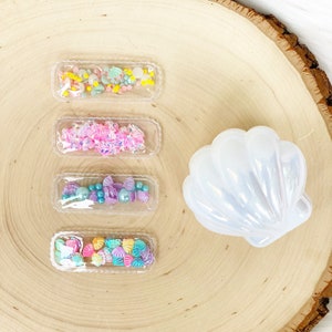 May include: Four clear plastic hair clips with different colourful embellishments inside, including seashells, beads, and glitter. A white seashell-shaped object is also in the image.