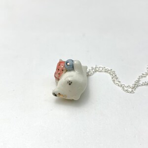 May include: White ceramic necklace featuring a small, detailed figurine of a cat carrying a strawberry and a blueberry. The necklace is on a silver chain.