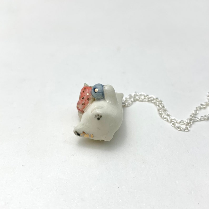 May include: White ceramic necklace featuring a small, detailed figurine of a cat carrying a strawberry and a blueberry. The necklace is on a silver chain.