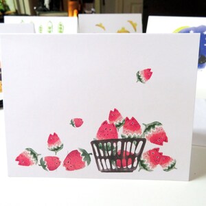 May include: A white card featuring a black wire basket overflowing with red strawberries. The strawberries have cute faces and are spilling out of the basket.