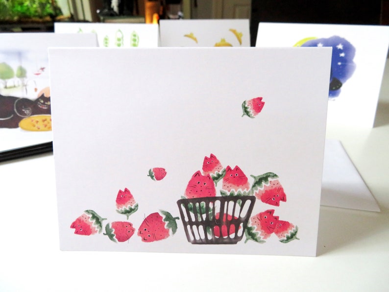 May include: A white card featuring a black wire basket overflowing with red strawberries. The strawberries have cute faces and are spilling out of the basket.