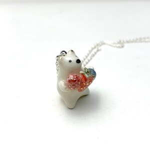 May include: White ceramic pendant necklace featuring a small bear holding two red strawberries. The necklace has a silver chain.