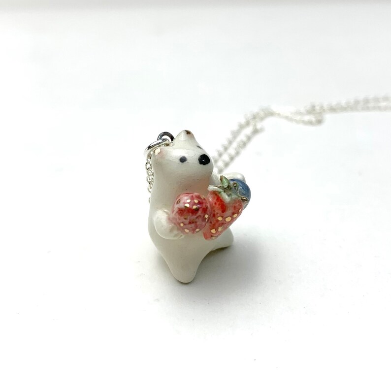 May include: White ceramic pendant necklace featuring a small bear holding two red strawberries. The necklace has a silver chain.