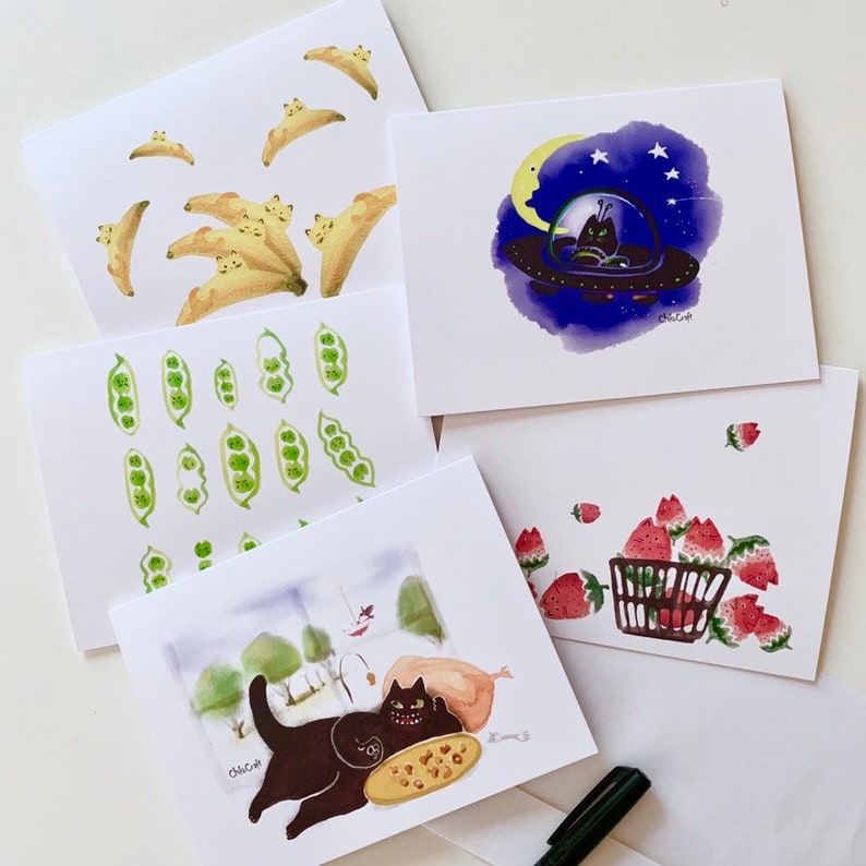 May include: Set of five illustrated greeting cards featuring various designs. One card shows a black cat in a spaceship, another shows a black cat with a pizza, another shows a basket of strawberries, another shows a row of green peas, and another shows a row of yellow bananas.