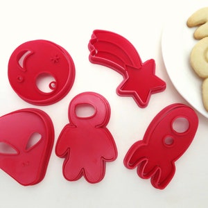 May include: Red plastic cookie cutters shaped like an alien, astronaut, rocket, shooting star, and a moon with a crescent moon and a star on it.