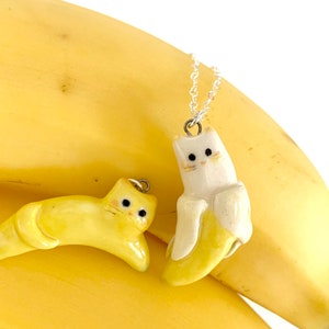 May include: Two ceramic cat pendants shaped like bananas. One is yellow and the other is white with a yellow banana inside. Both have black eyes and a silver chain.