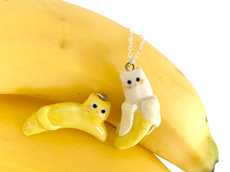 May include: Two ceramic cat pendants shaped like bananas. One is yellow and the other is white with a yellow banana inside. Both have black eyes and a silver chain.