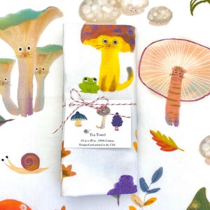 Op de afbeelding: Witte theedoek met een grillig bosontwerp met paddenstoelen, een kat, een kikker en een slak. De tekst "Tea Towel 19 in x 28 in. 100% Cotton Designed and printed in the USA" is op de theedoek gedrukt.